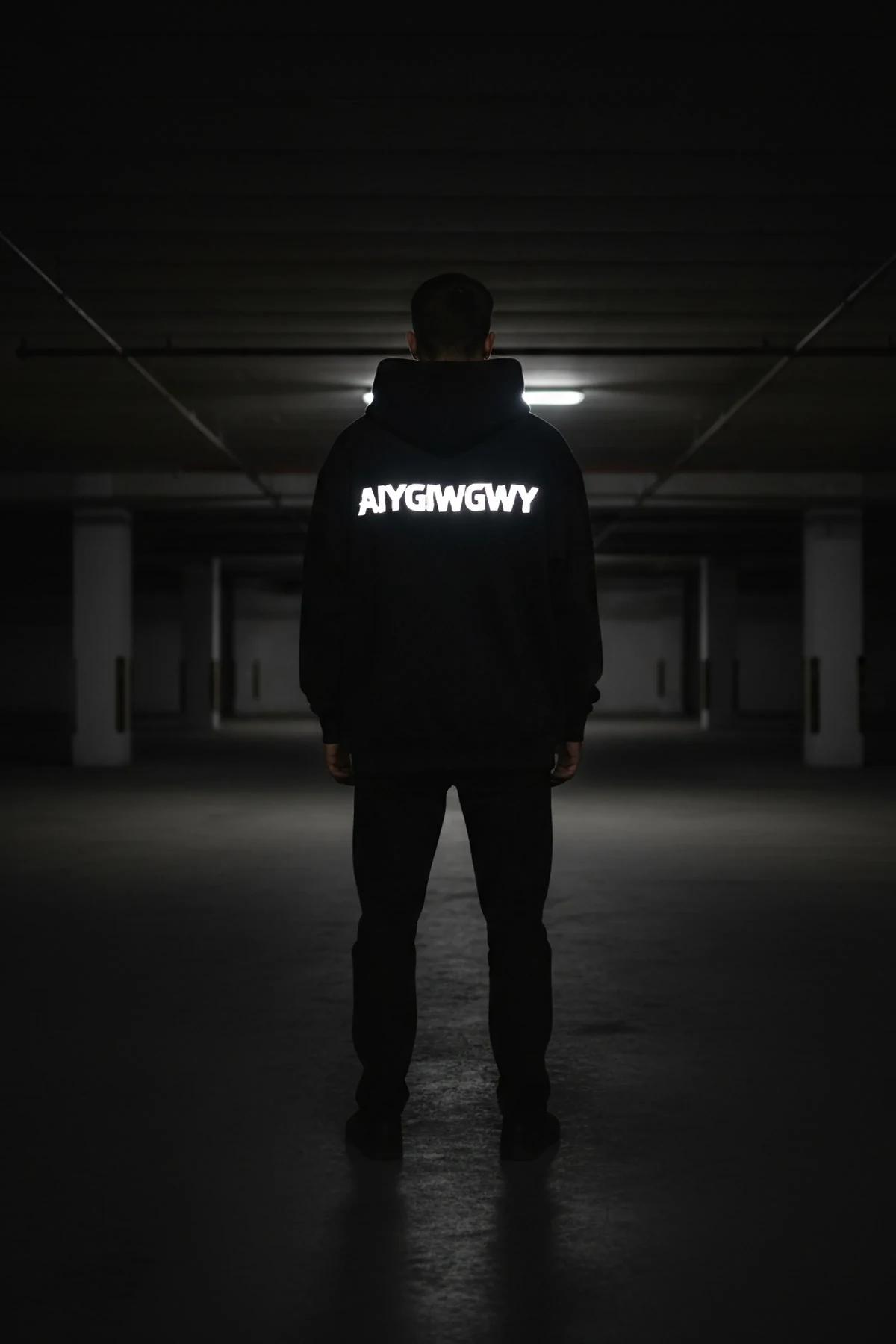 Black glow-in-the-dark hoodie with bold white reflective custom text on chest, worn in dark urban parking garage