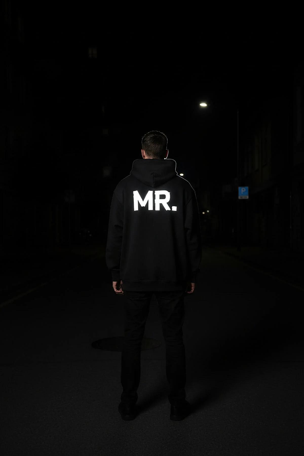back view of black oversized hoodie with reflective MRS. sleeve lettering glowing under streetlight