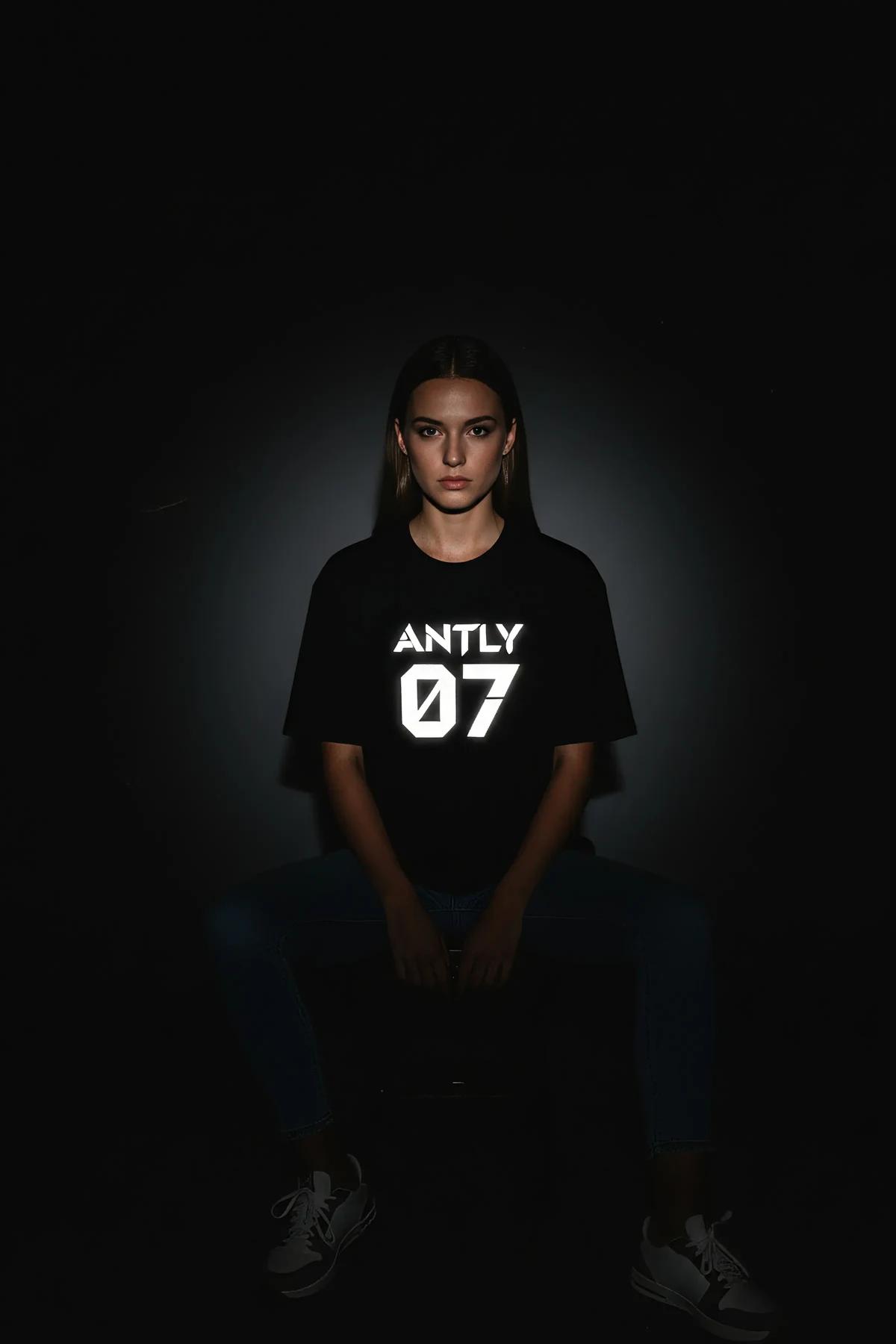 black oversized reflective graphic t-shirt with glowing white ANTLY 07 logo worn by woman in low light