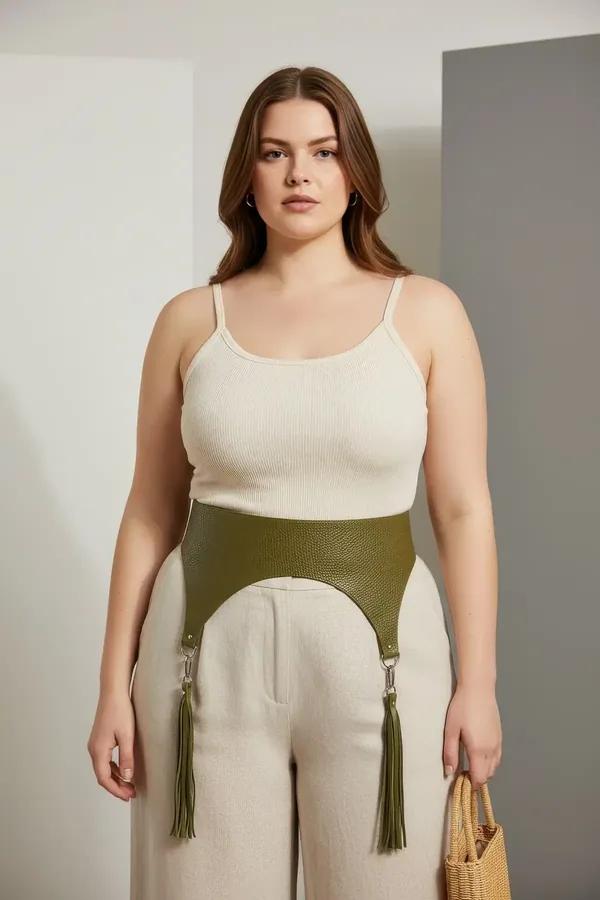 olive green high-waisted underbust harness belt with silver O-ring hardware and matching tassel garters worn over linen trous