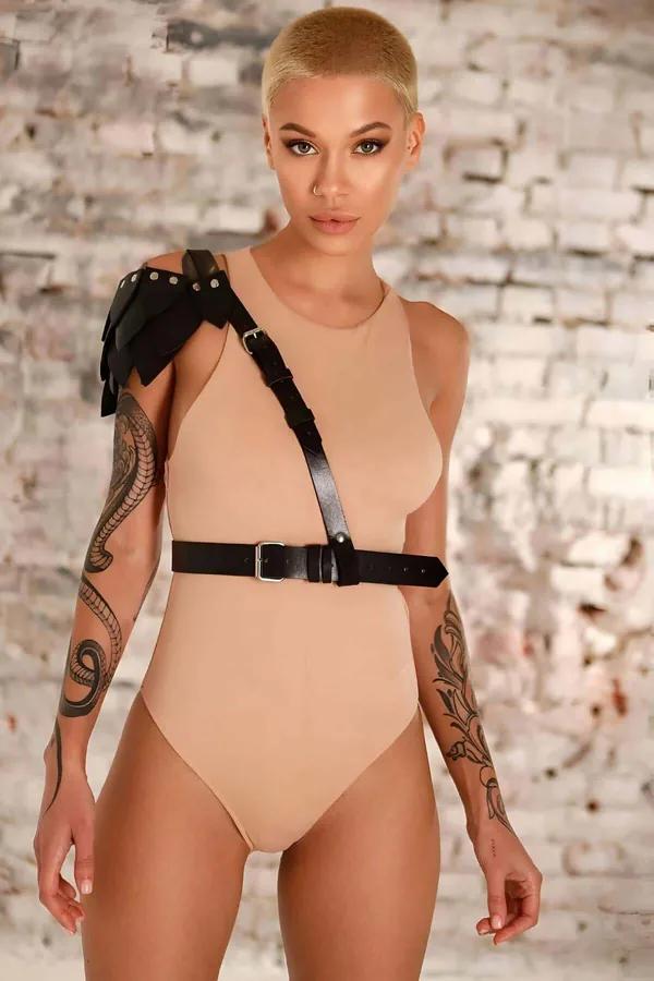 Black faux leather asymmetrical shoulder harness with stud epaulette and wide silver-buckle waist belt worn over beige bodysu