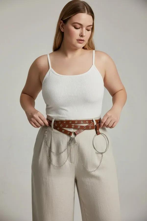 Brown faux leather waist harness belt with silver grommets draped chains and O-ring worn over white top and cream maxi skirt