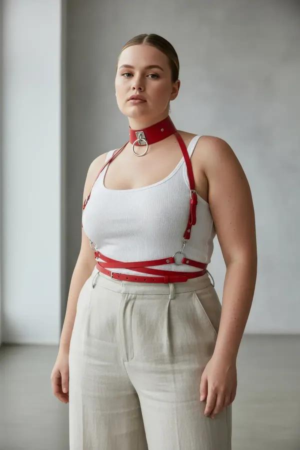 red faux leather harness bra with silver O-ring choker and waist belt worn over cream tank top, plus-size model
